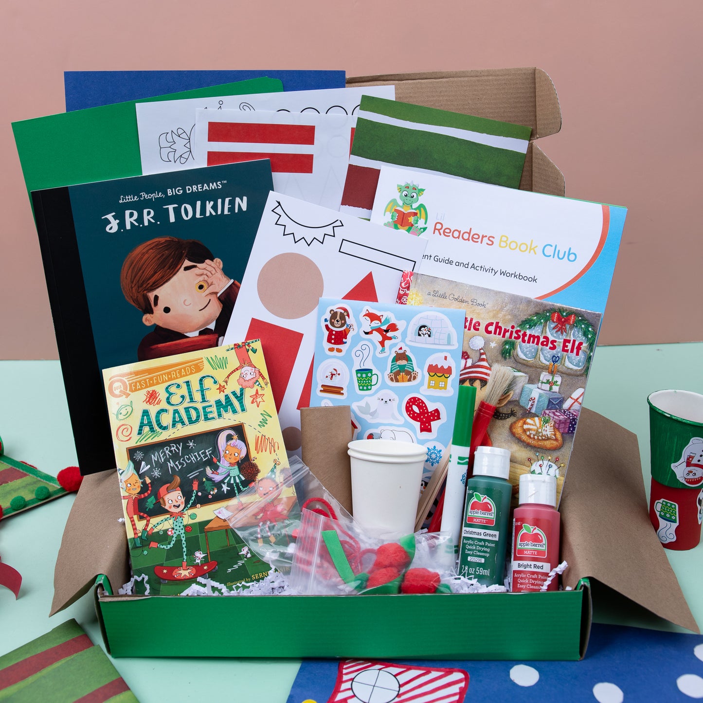 A book club box with themed crafts and materials for kids, including a book titled 'Elf Academy', a set of stickers, and a craft kit, all presented on a table with a Christmas tree in the background.